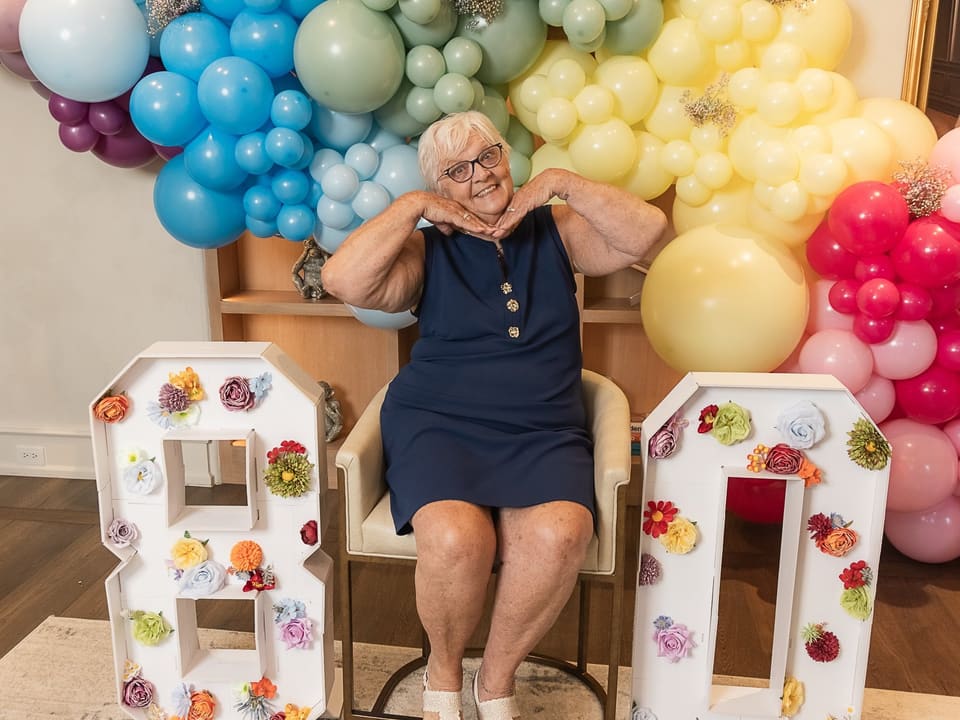 Guest of honor smiling in a chair between floral 8 and 0 letters under a balloon arch