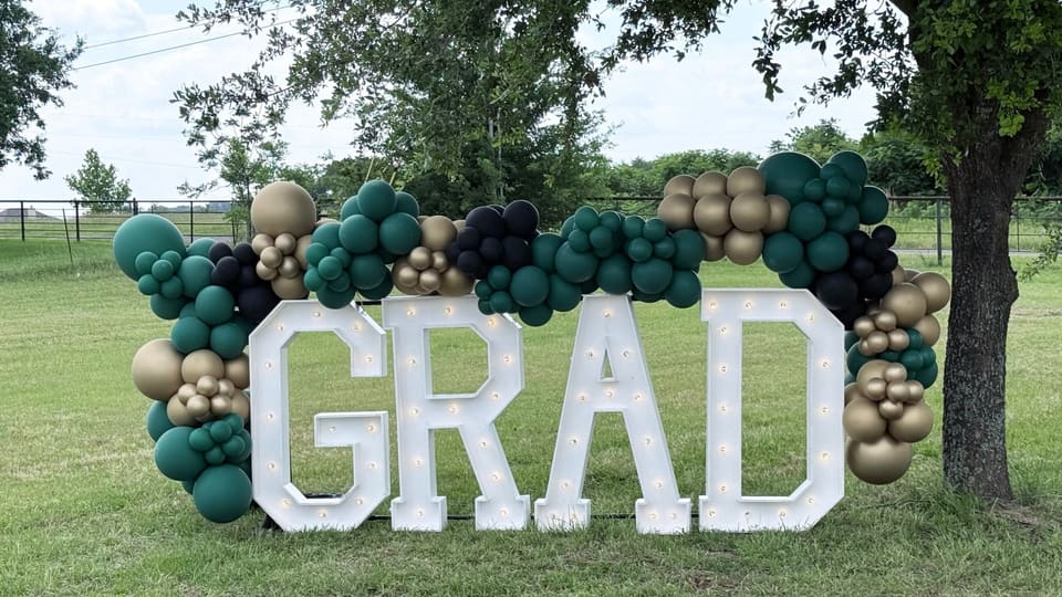 Lighted GRAD marquee letters framed by green and gold balloons in a field